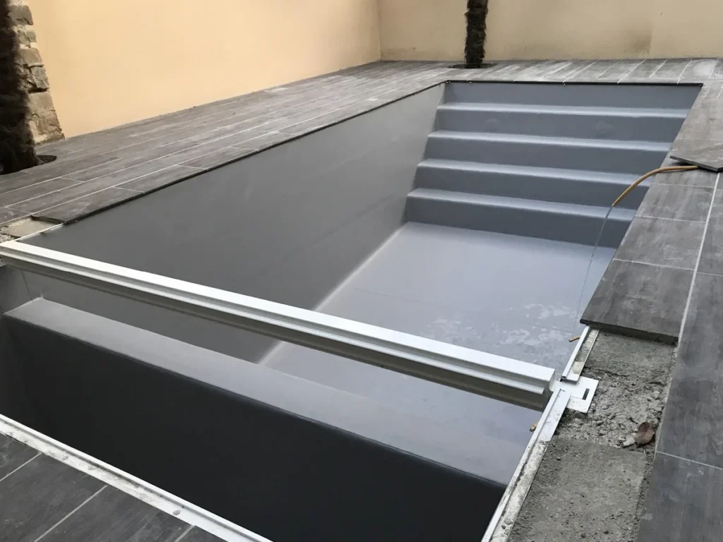 La même piscine après rénovation : membrane PVC armé gris clair, escalier d'angle et plage en carrelage imitation bois